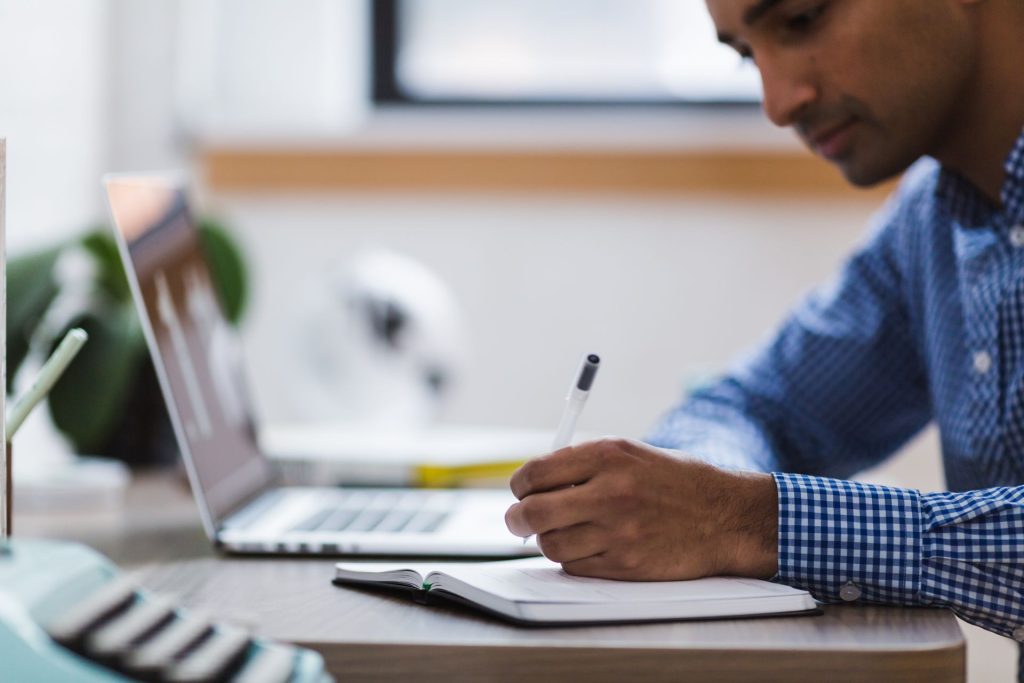 homme qui écrit sur papier avec ordinateur portable a coté 