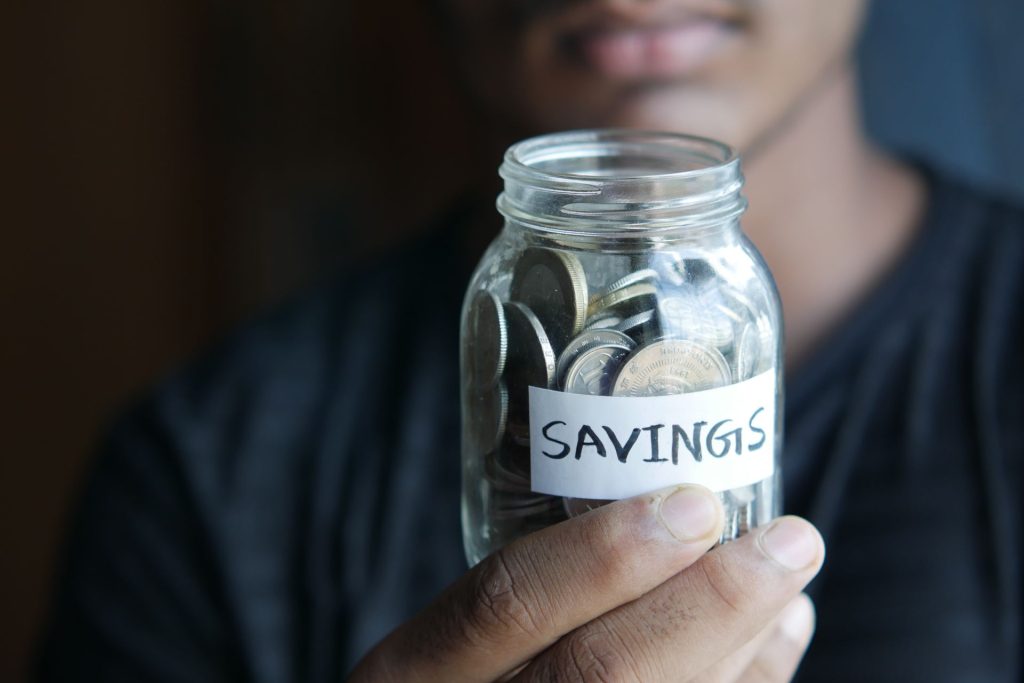 homme tenant un bocal remplit de pièces de monnaies avec ecrit savings dessus 